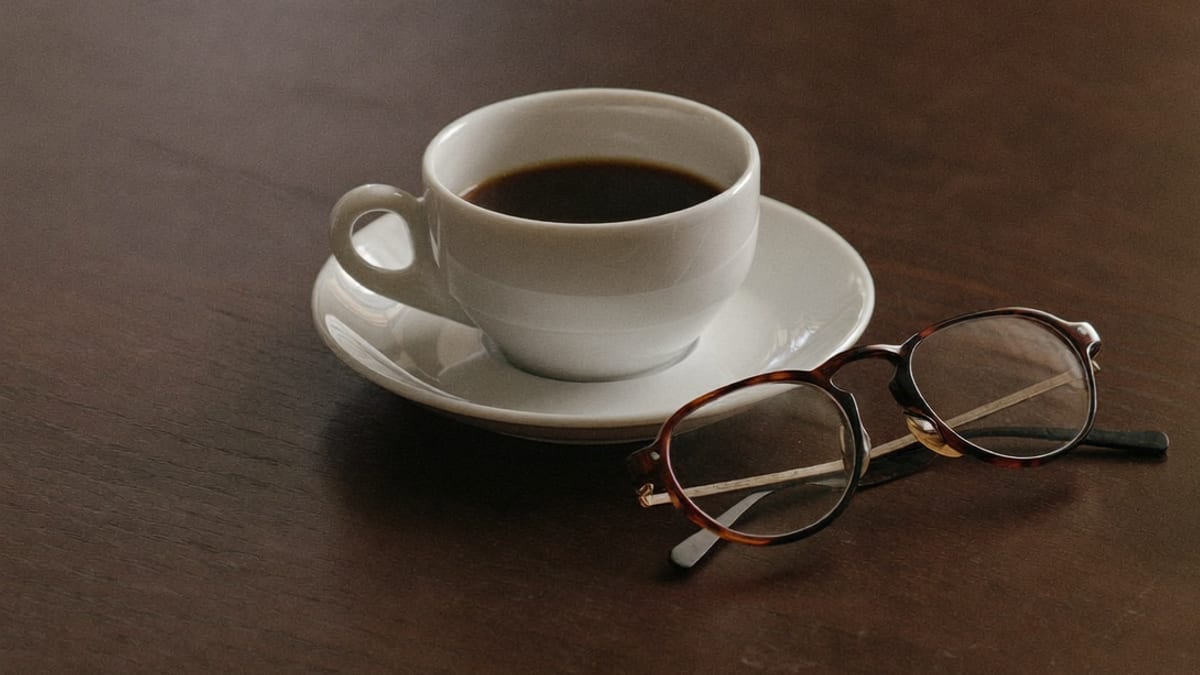 A cup of coffee and reading glasses on a dark wooden table